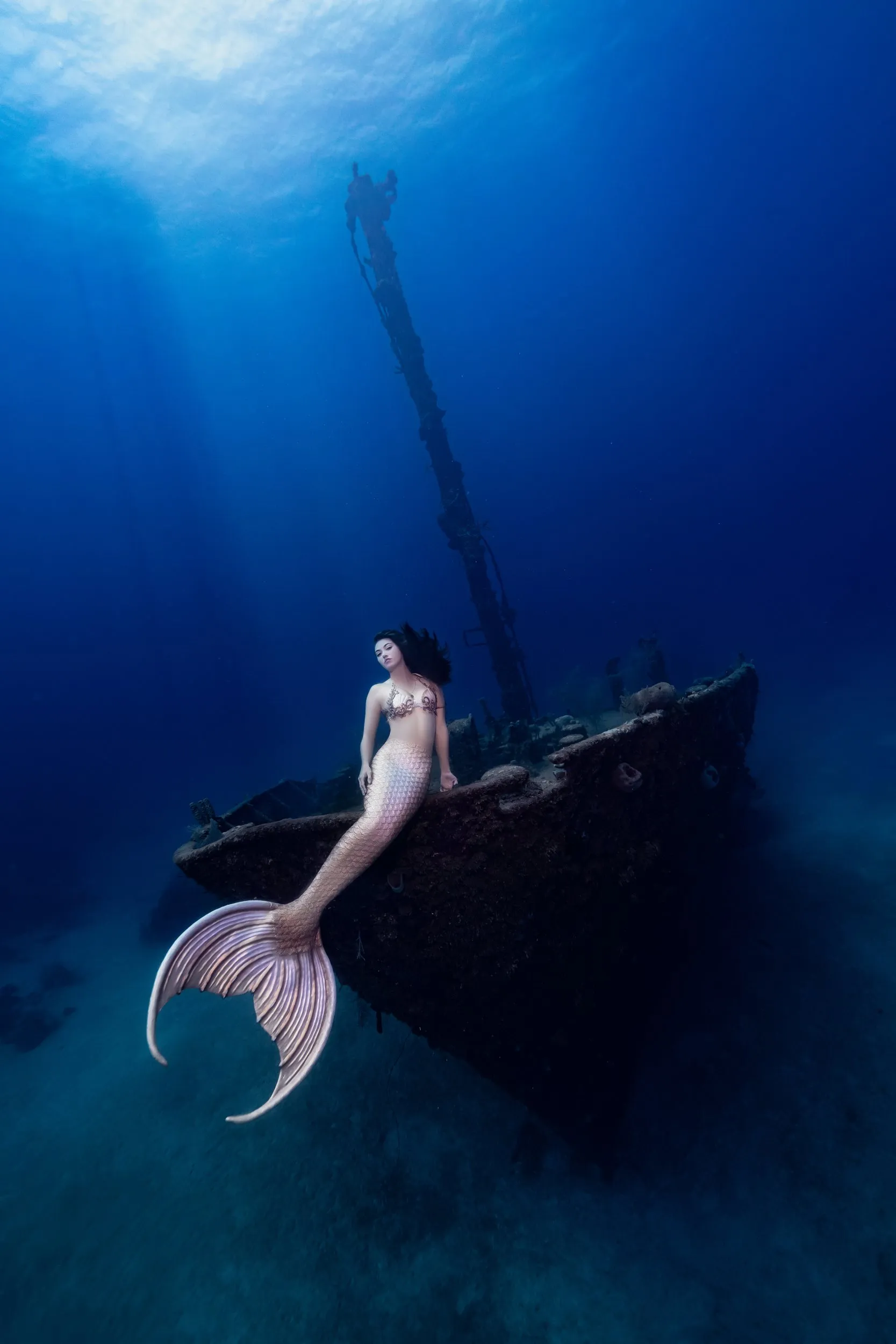 A mermaid with a pale pink tail rests on the bow of a sunken ship in deep blue water, sunbeams falling from above.