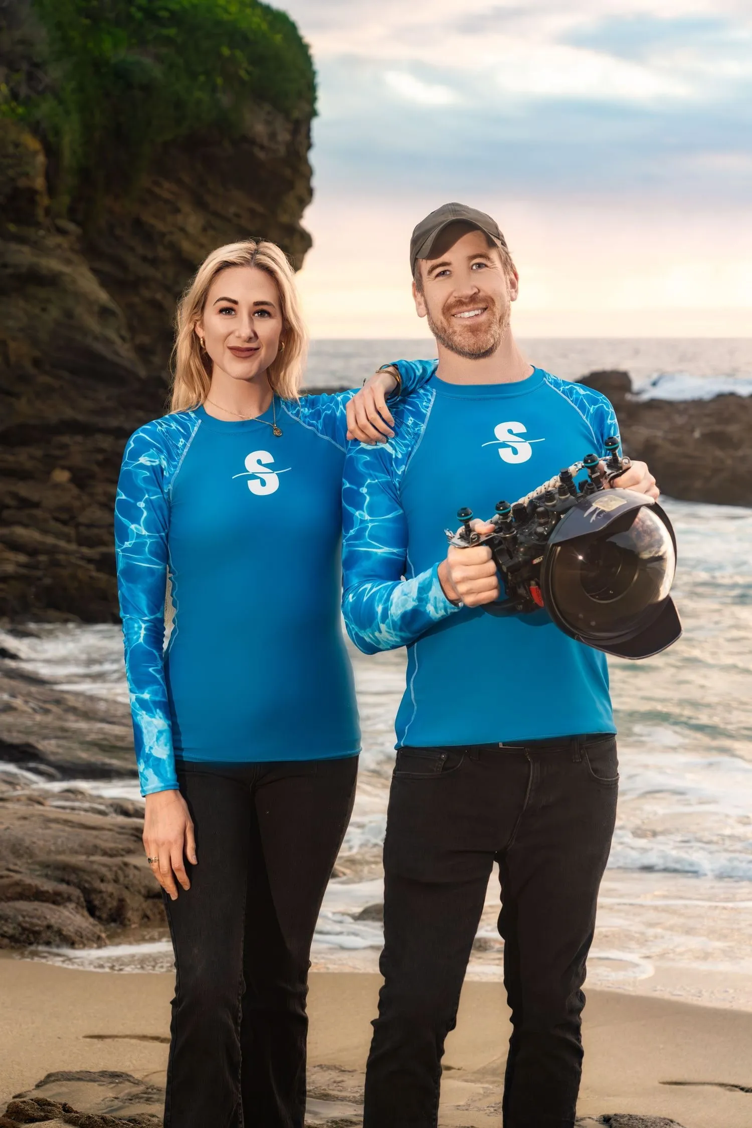 Justin and Abbey standing on the beach in Submerge rash guards, Justin holding an underwater camera housing.