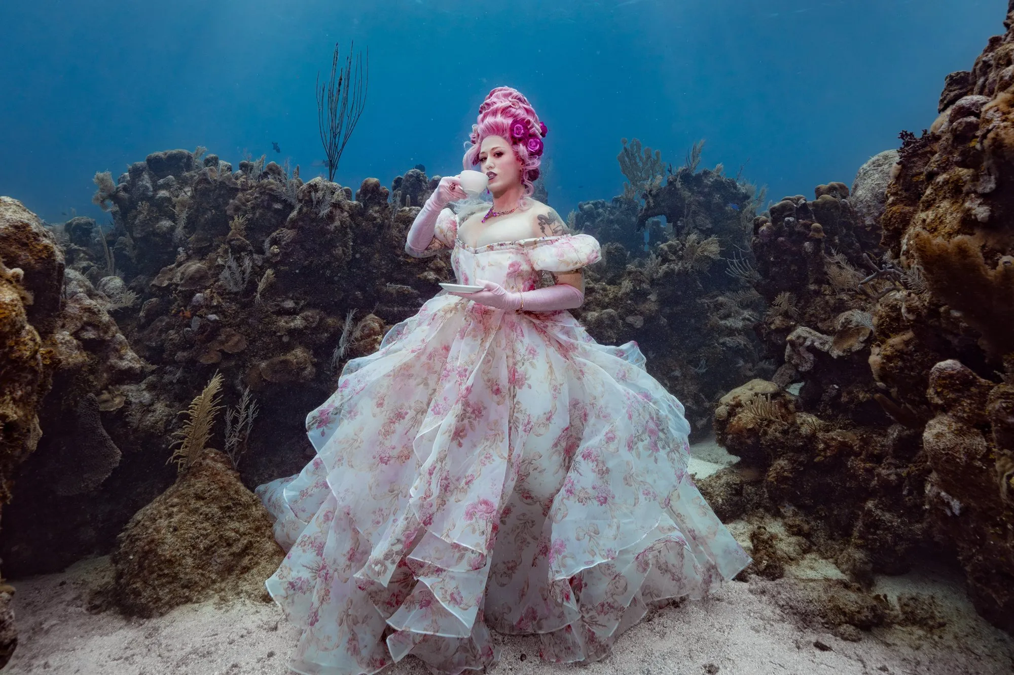 A woman with a pink powdered wig and a floral organza gown sips tea from a porcelain cup and saucer on a coral reef, sunbeams falling through blue water behind her.