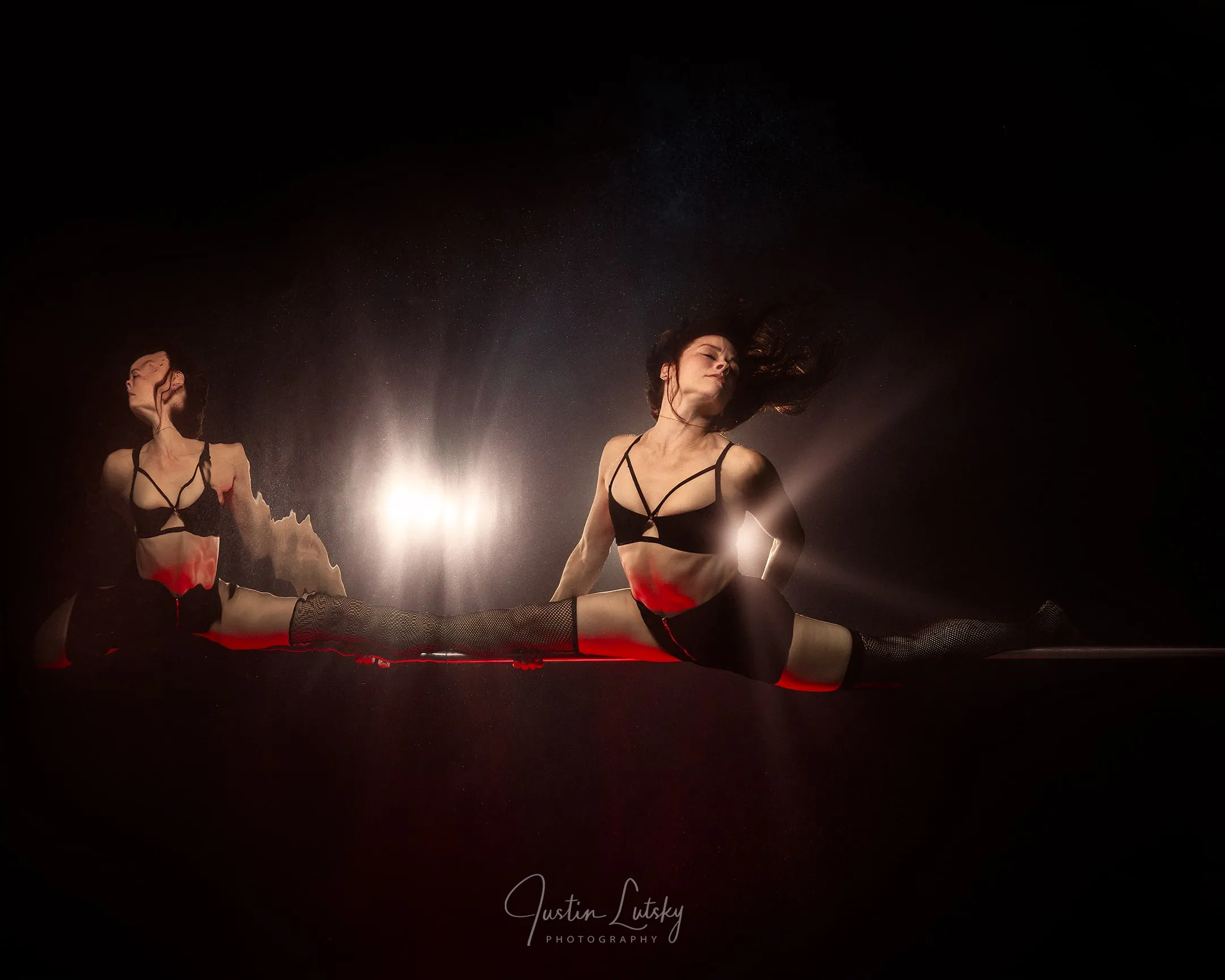 A performer in black fishnets holds a full split on a pole at the waterline, mirrored in the surface above, backlit by a single warm beam through dark water.