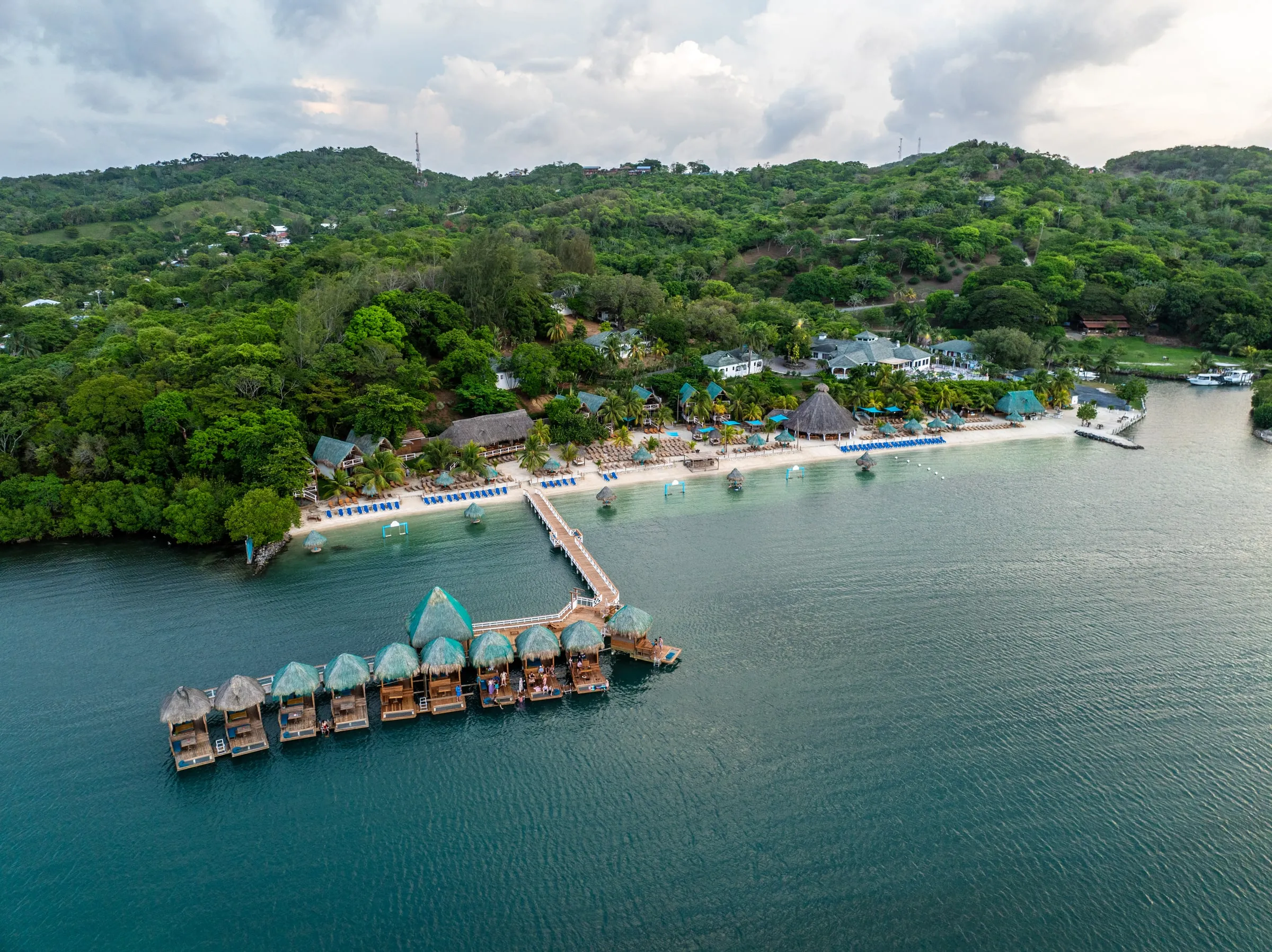 Turquoise Bay Resort from the air: a row of thatched overwater cabanas at the end of a wooden pier, the beach and main buildings along the shore, green hills rising behind.