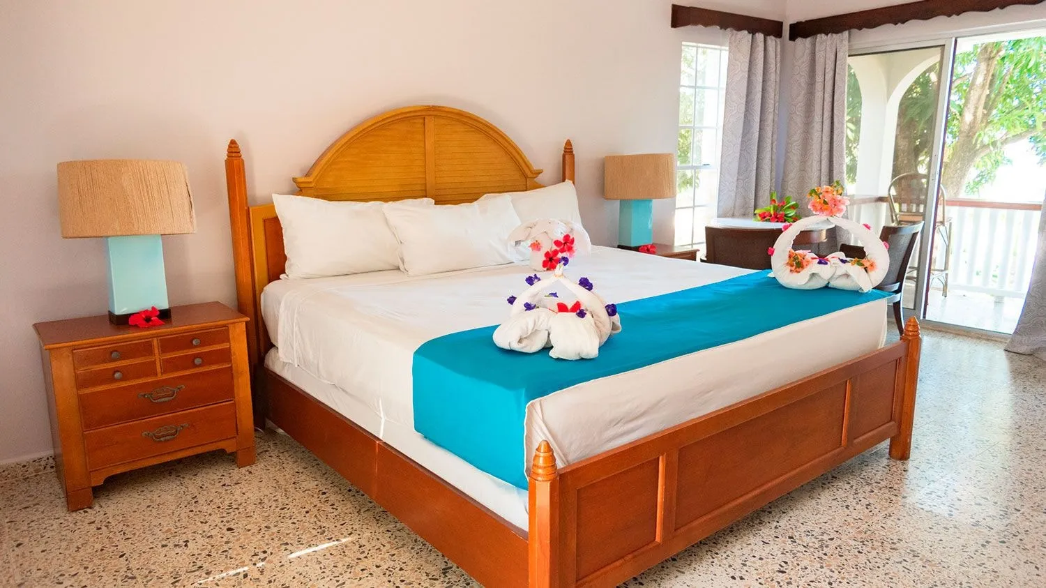 A king bed with a turquoise runner and towel animals, a balcony door open to the garden.