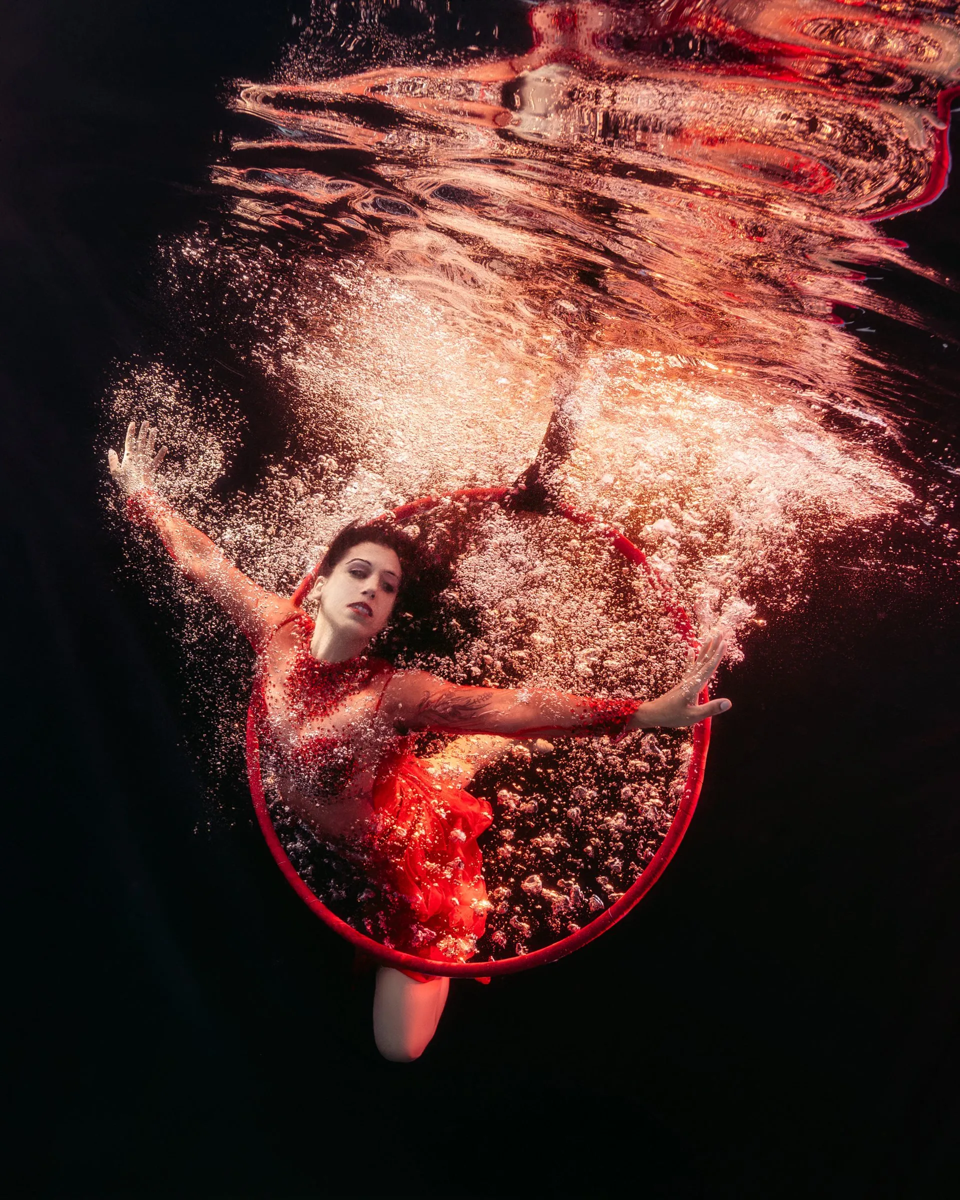 A performer in a red costume curls inside a red hoop, her hair floating, in a spray of bubbles lit orange and red.