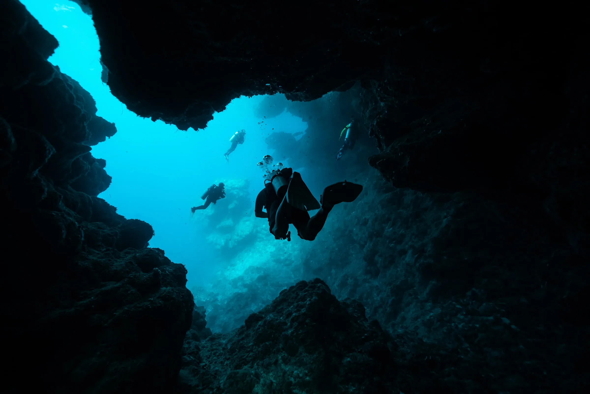 A scuba diver swims through a dark cave opening toward blue water, sunlight on the rock around the arch.