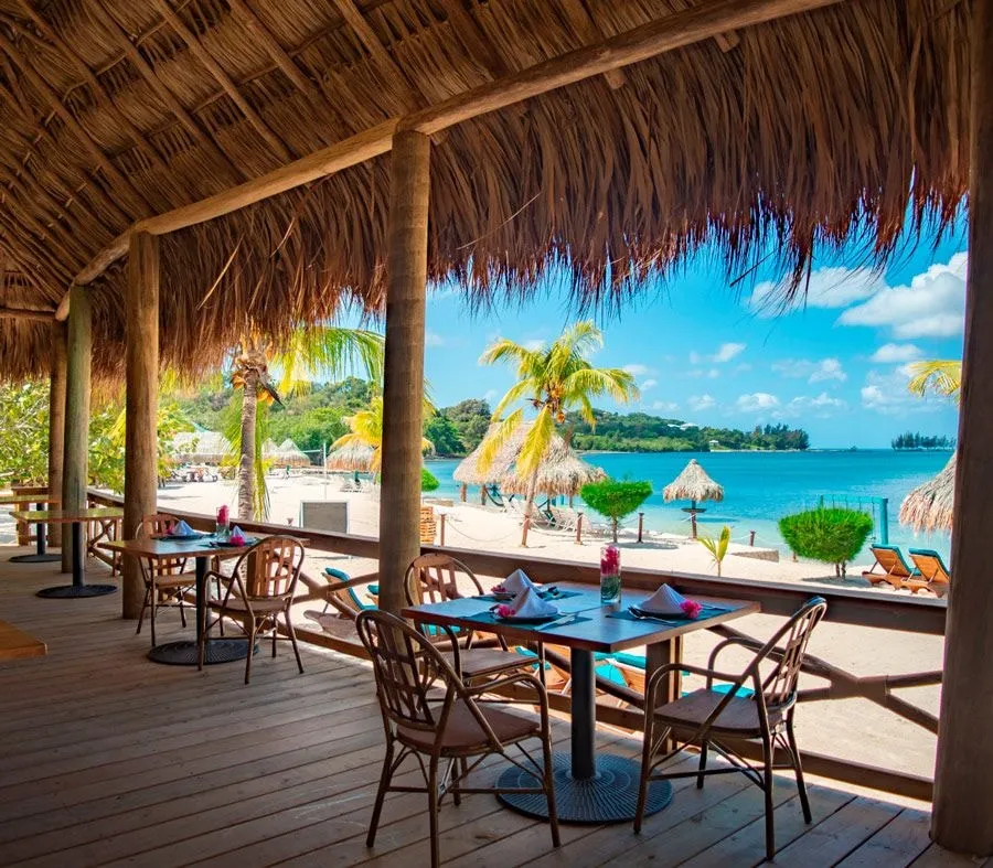 A thatched-roof restaurant deck with set tables looking out to the beach and palapas.
