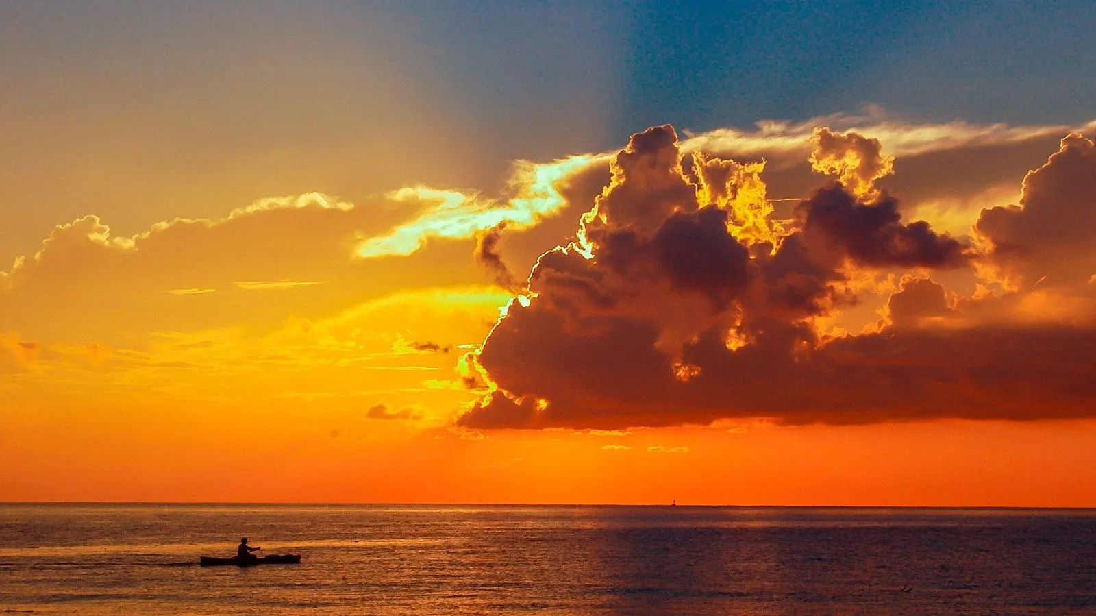 A kayaker paddles beneath an orange sunset sky over the sea, backlit clouds above the horizon.