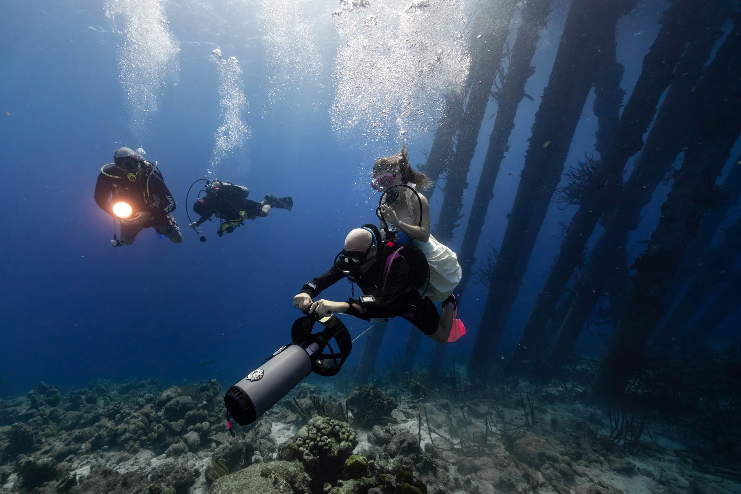 A diver rides an underwater scooter with a woman in a white gown holding on behind him, two divers with a light following among the pier pilings.