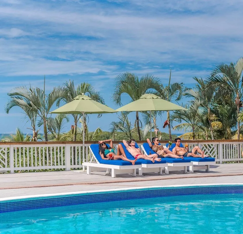 Guests sunbathing on blue loungers beside the pool, palms and green umbrellas behind them.