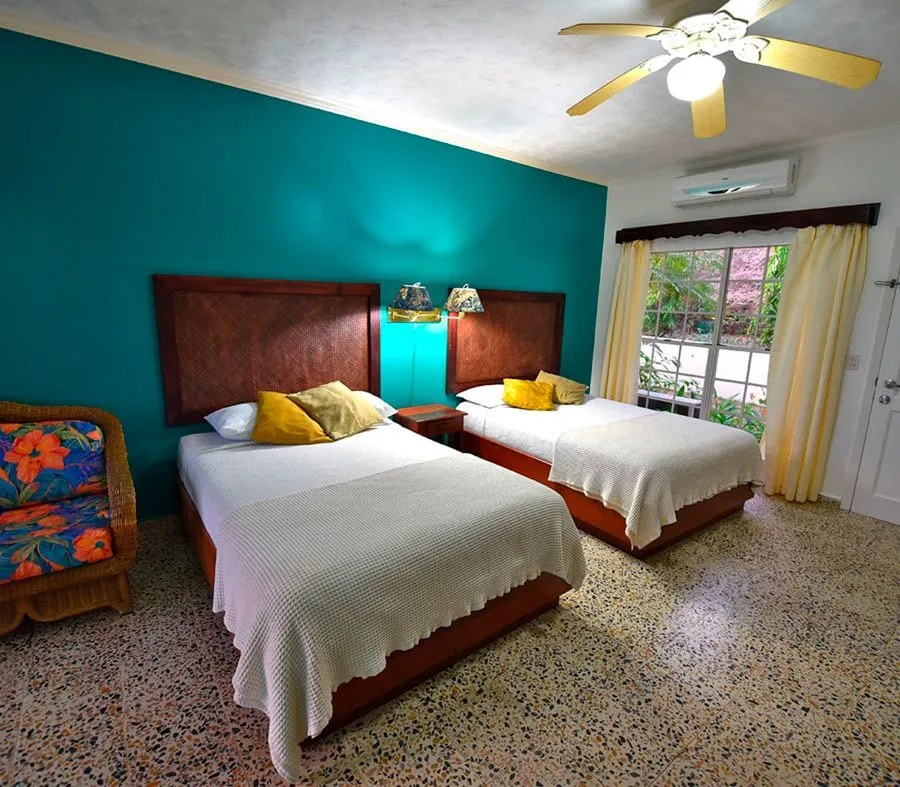 A Hillside room: two beds under a ceiling fan, a teal wall and a bright patterned chair.