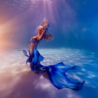 A mermaid in a blue tail rises through bright water, colored rays of light falling across a sunlit pool floor.