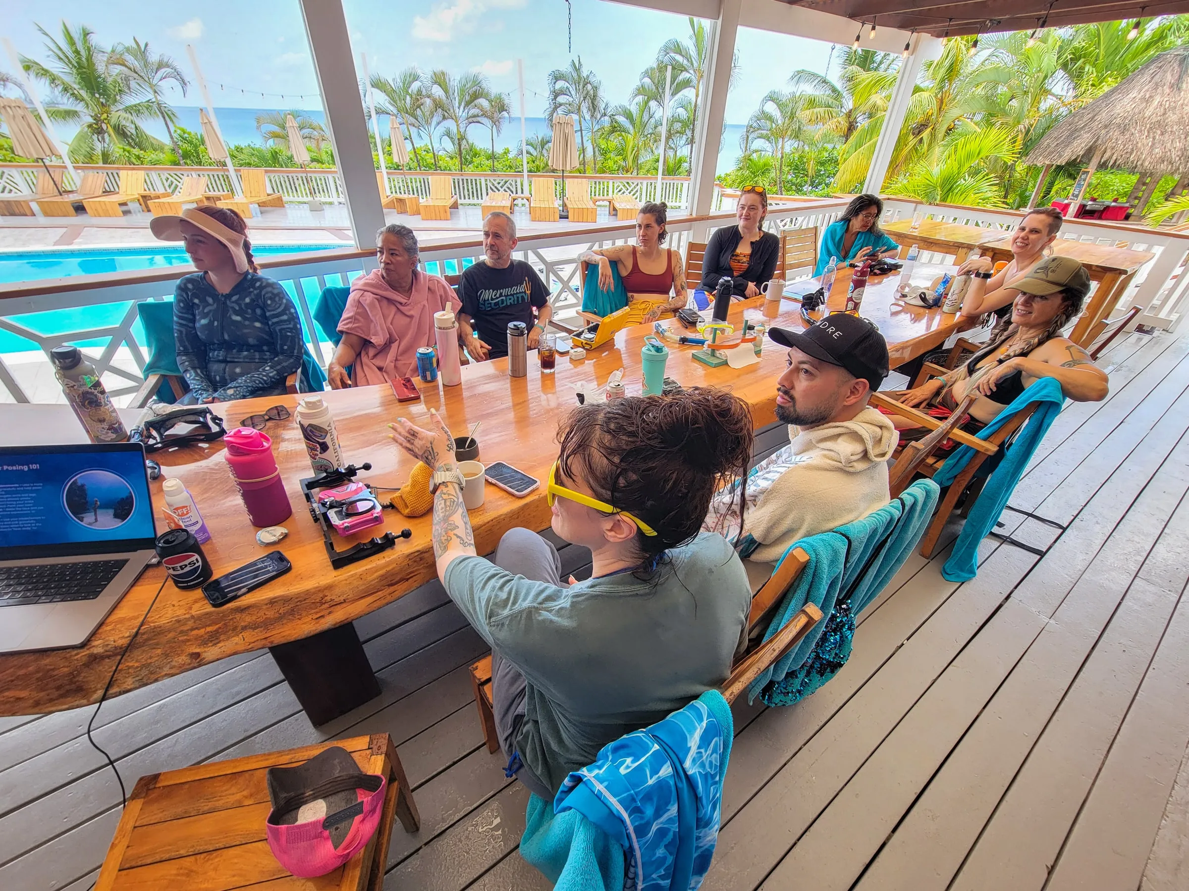Workshop guests around a long wooden table on the resort deck, one of them gesturing as a posing slide shows on a laptop, the pool and the sea behind.