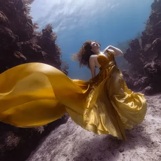 A woman in a gold gown with a long trailing skirt stands on a sandy ledge between two reef walls, light shimmering through the water above.