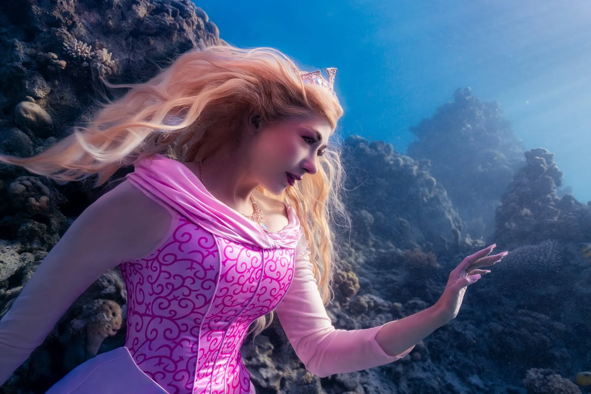 A woman with long blonde hair in a pink princess gown and a small tiara reaches one hand out beside a coral reef, blue water and sunbeams behind her.
