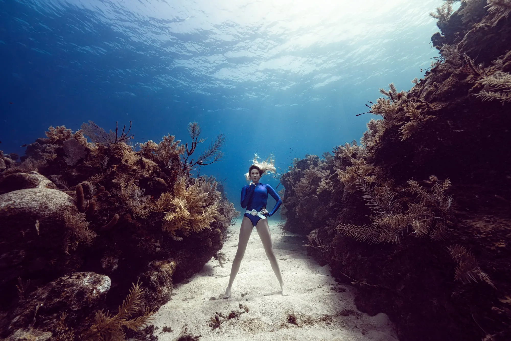 A freediver in a blue suit and weight belt stands barefoot on white sand between two reef walls, one hand at her collar and her hair floating up toward the sunlit surface.