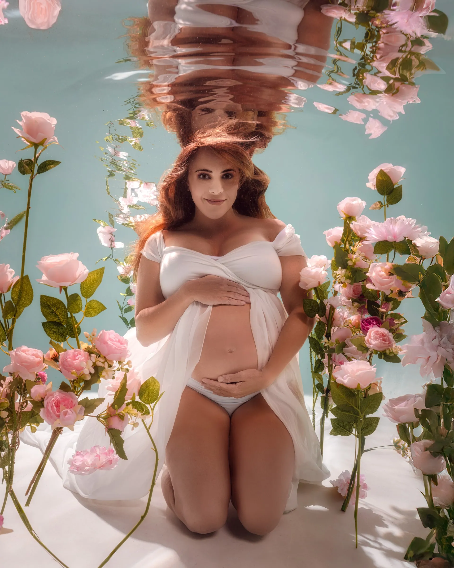 An expectant mother in a white top and wrap kneels among pink roses against pale blue, both hands resting on her belly, her reflection hanging above her.