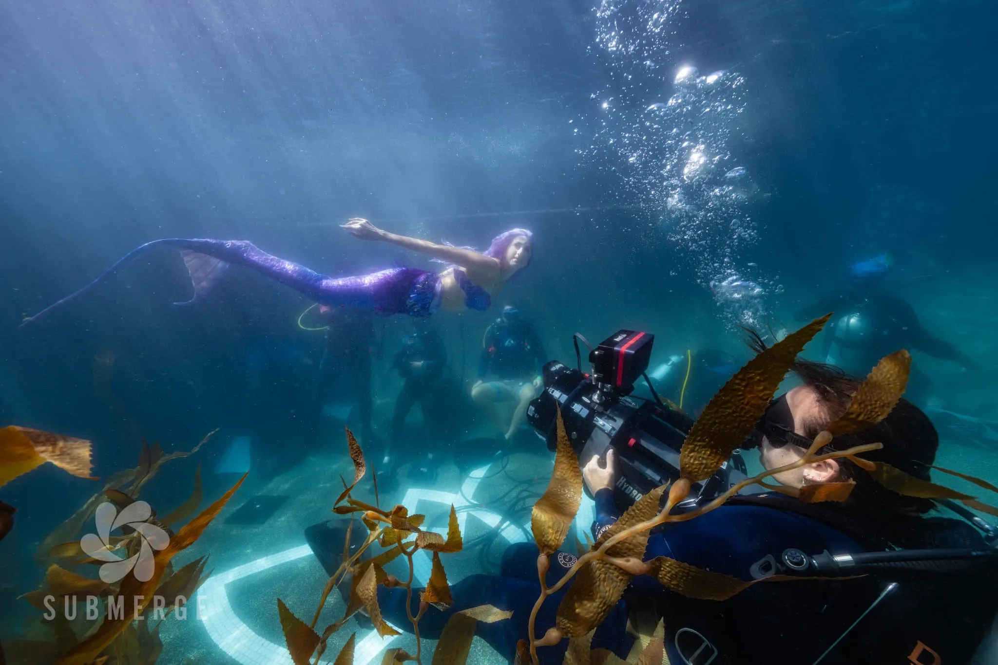 An operator in a blue wetsuit films a mermaid with purple hair swimming above a glowing ring on the pool floor, bubbles rising and kelp across the foreground.