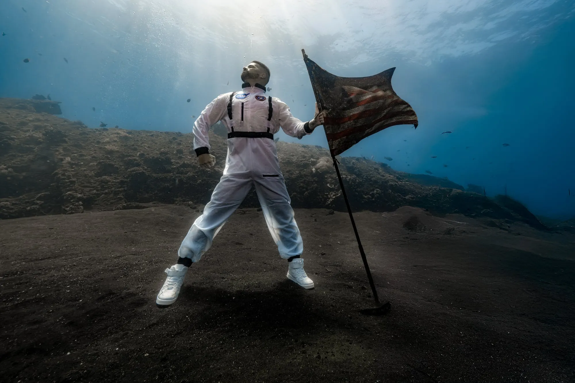 A man in a white spacesuit stands with his feet apart on black sand, holding a flag on a pole, sunbeams in the blue water behind him.