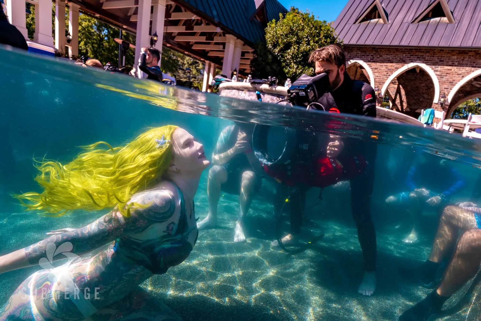 A performer with long yellow hair floats at the surface of a pool while crew watch and a camera operator films from the deck.