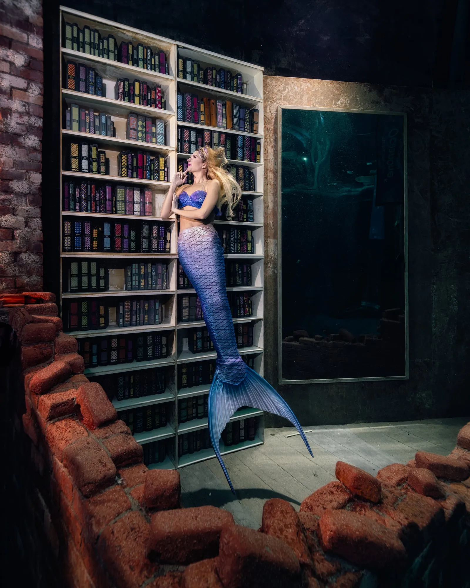 A mermaid with a purple tail hangs in front of a tall bookshelf in a ruined brick room, one hand at her chin.