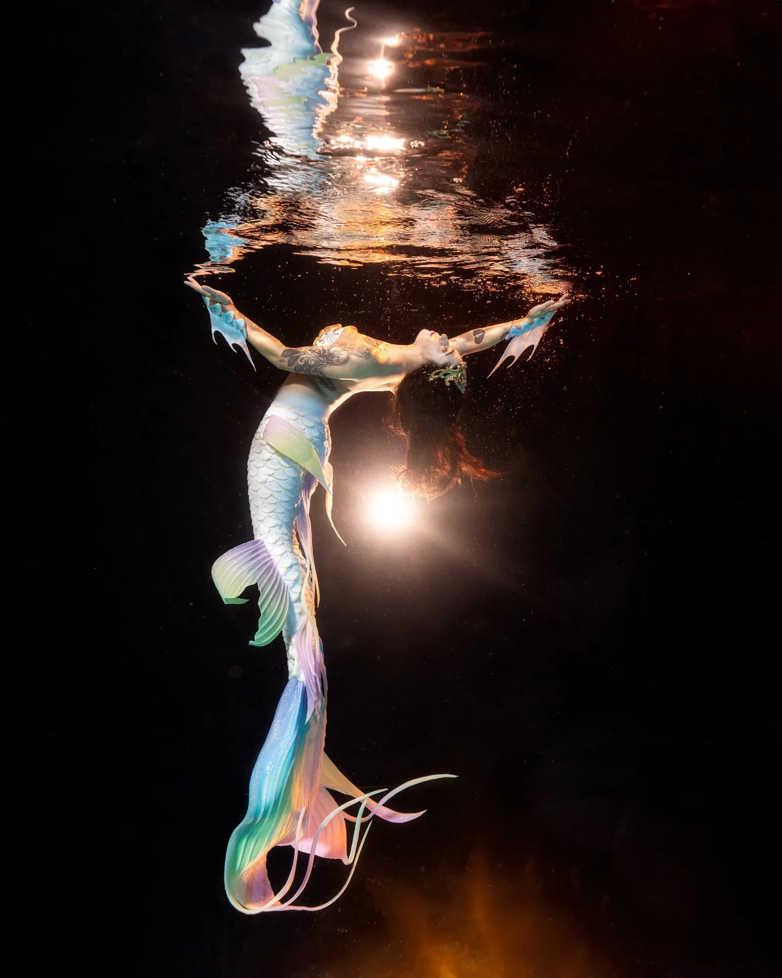 A mermaid in a pearl and pastel tail floats arms out beneath the surface with a light glowing behind her, in black water.