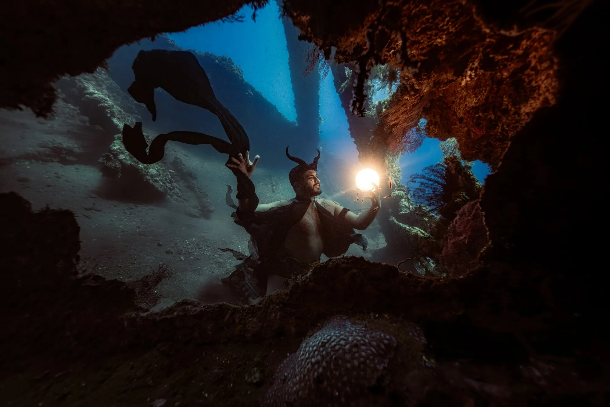 A figure with curved horns and a black cape holds a glowing orb inside a shipwreck, an opening in the rusted hull lit blue behind.