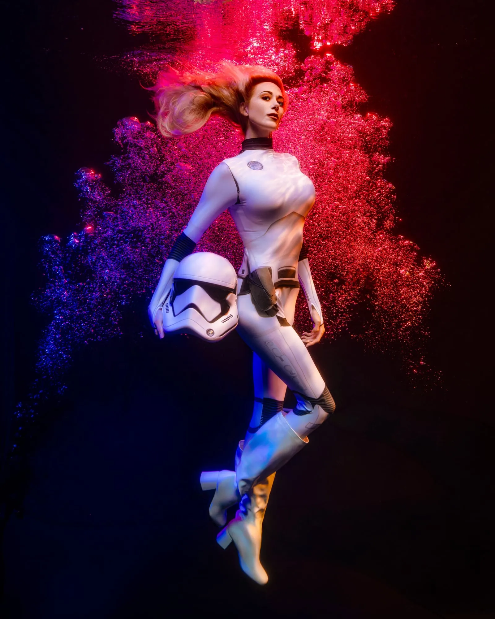 A woman in a white storm-trooper costume and white boots holds a helmet at her side as she floats upright in a burst of red and violet bubbles on black water.