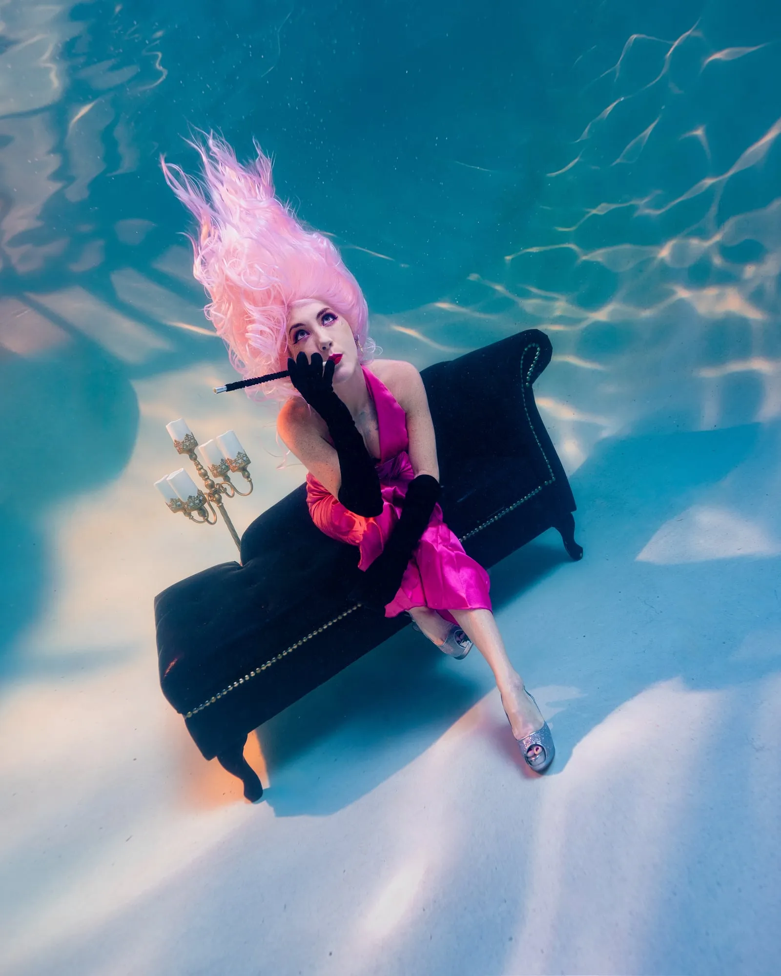 A woman with tall pink hair and a hot pink dress rests her chin on a black glove, seated on a black chaise lounge beside a candelabra, seen from above.