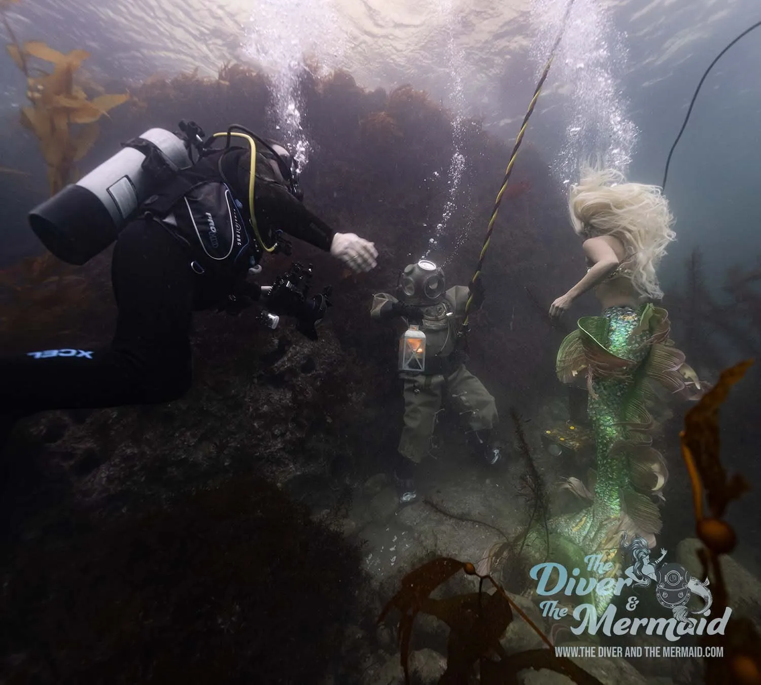 A camera operator in scuba gear films a diver in a brass helmet and a mermaid with long blonde hair among kelp and rock.