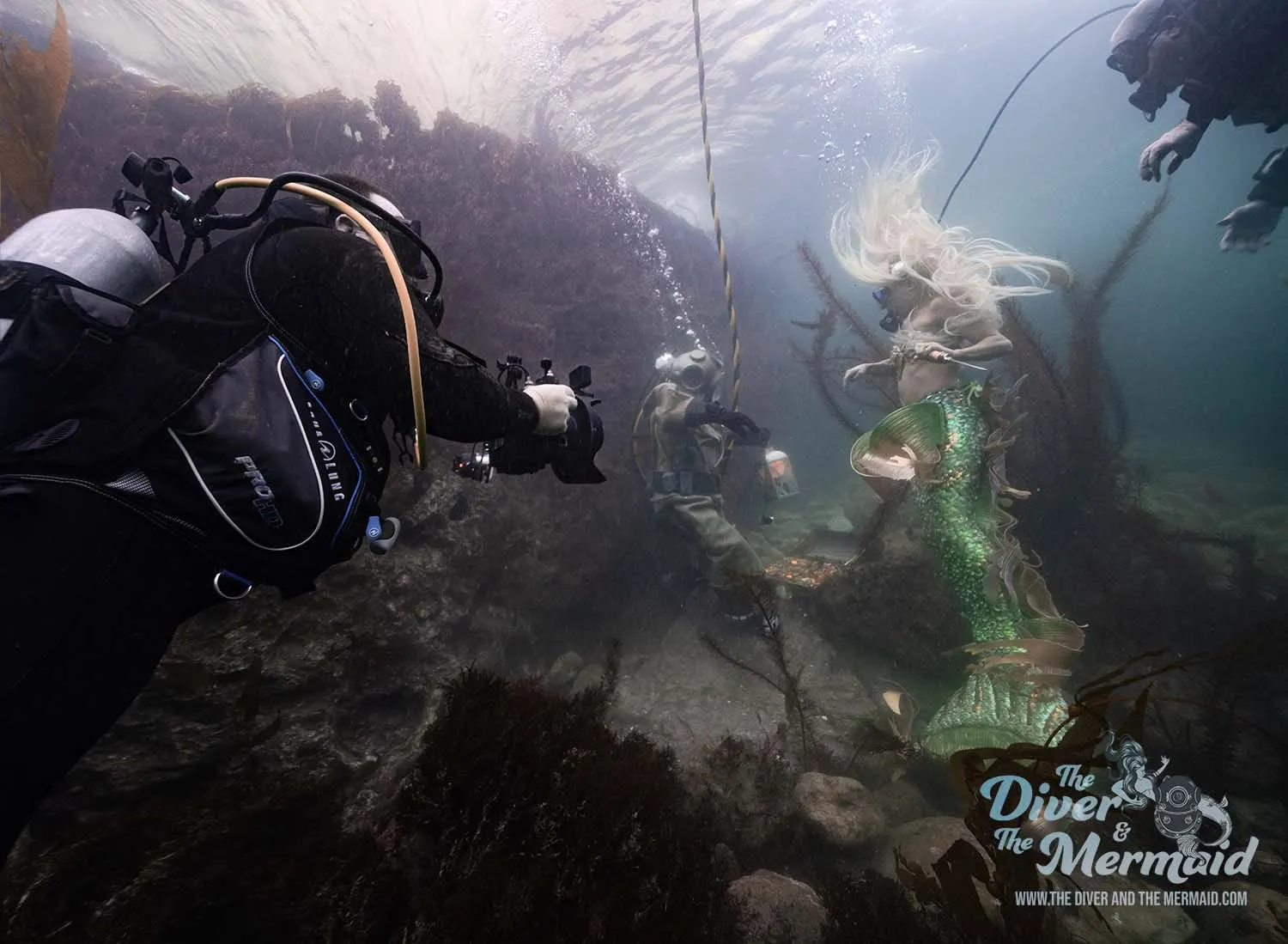 From behind an operator in scuba gear, a diver in a brass helmet and a mermaid in a green tail stand among kelp and rock.