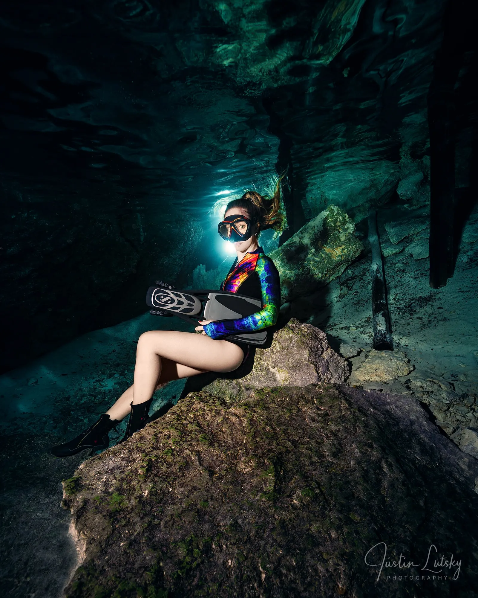 A freediver in a rainbow top sits on a rock in a dark cave, a torch lighting her face and long fins in her hand.