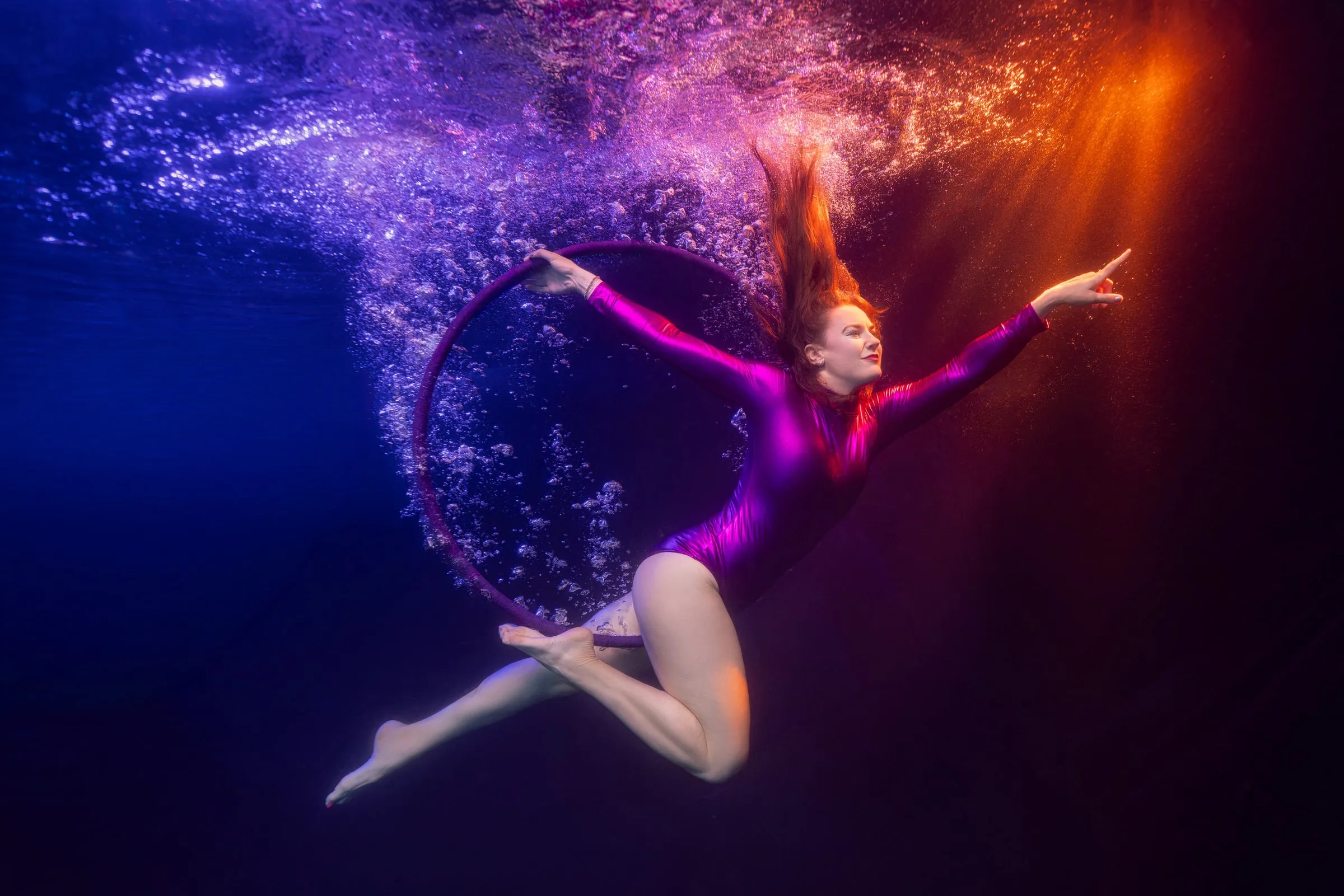 A performer in a purple leotard arches away from a hoop with one arm pointing out, her red hair streaming upward through blue water with orange light behind her.