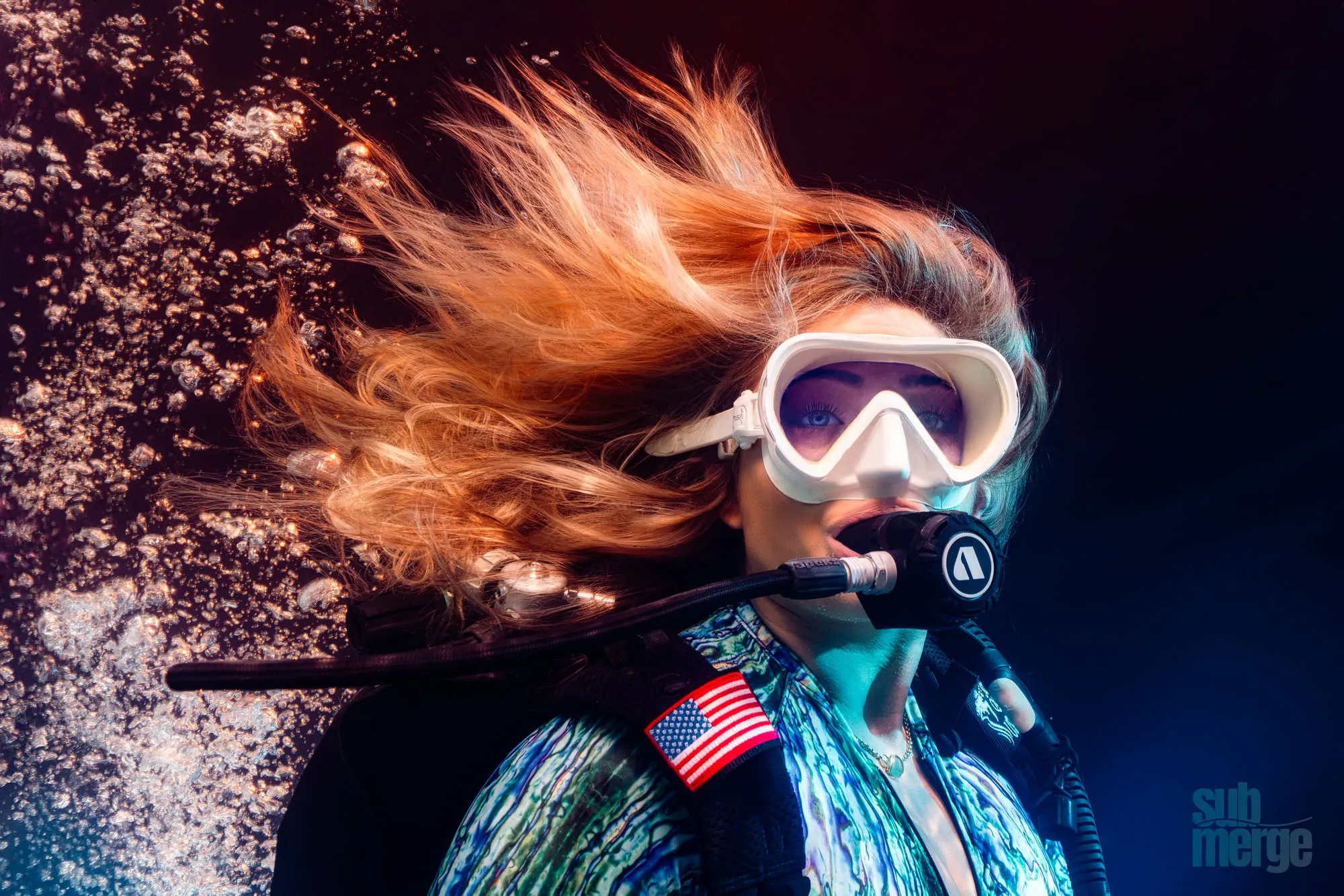 A diver with orange hair streaming and a US flag on her jacket breathes from a regulator in dark water among bubbles.