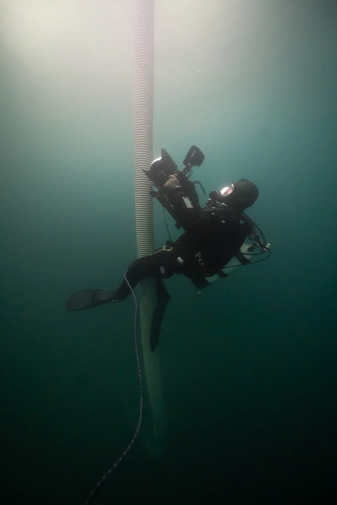 A diver with a camera and a light climbs a pole in dark green water, a bright light above him.