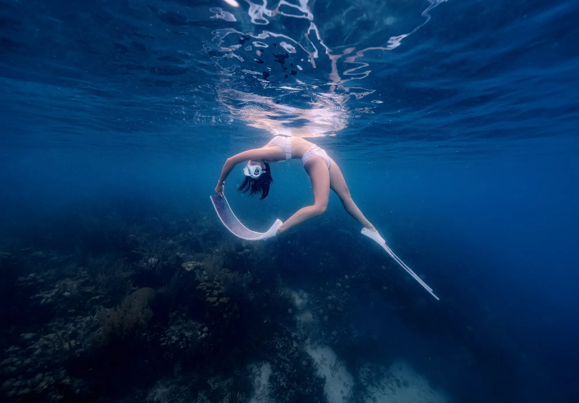 A freediver in a white swimsuit arches backward just under the surface of deep blue water, her mask and long fins trailing, a reef far below.
