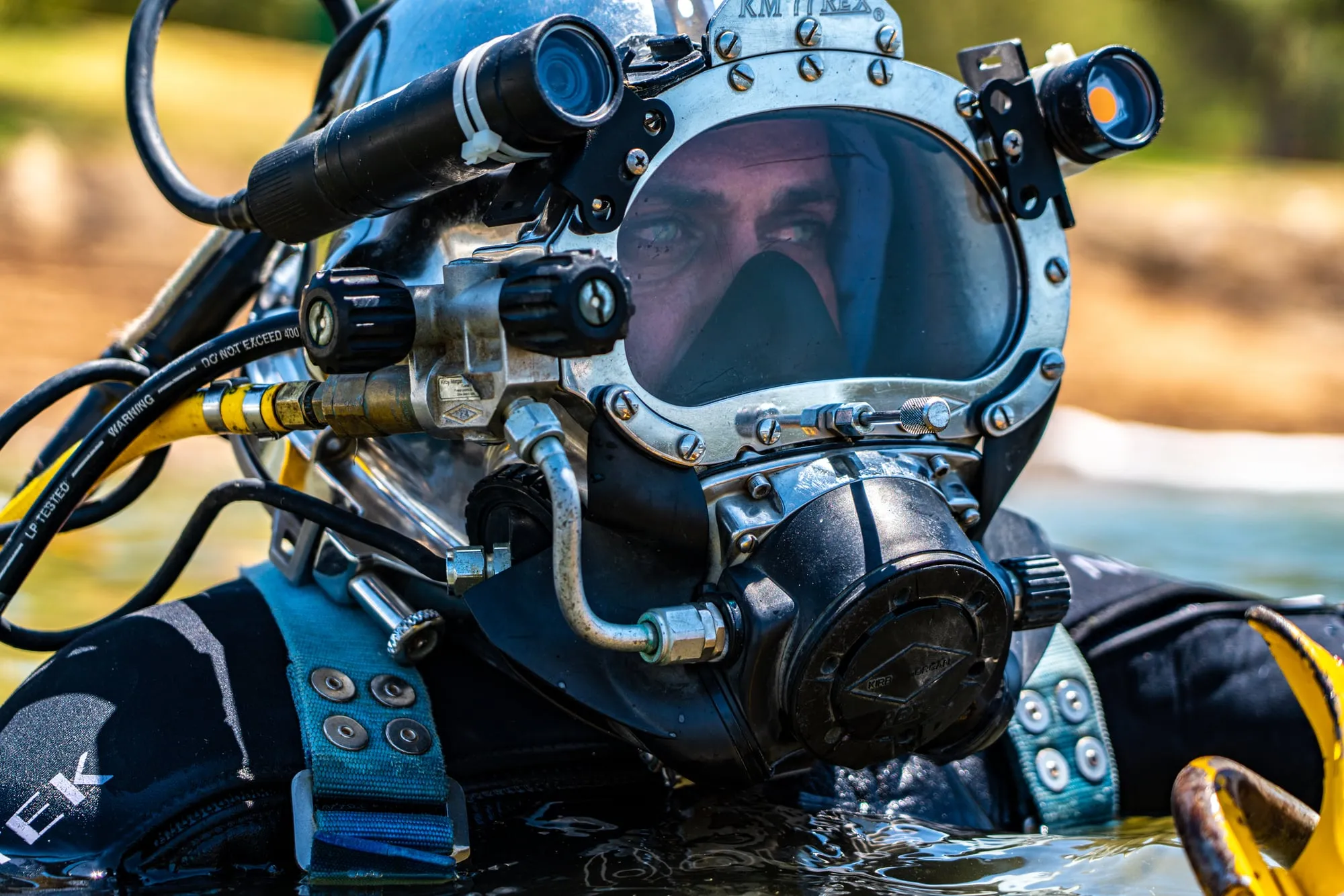 A commercial diver in a chrome helmet and black dry suit surfaces in a lake, hoses and a yellow harness around him.
