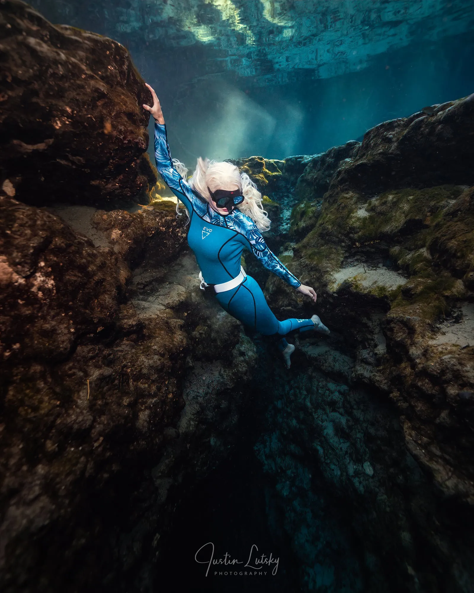 A freediver in a blue wetsuit climbs a rock face in a spring, one arm stretched up the stone and a mask on her face.