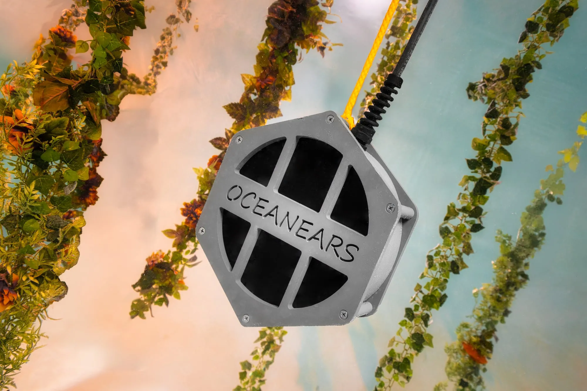 A hexagonal Oceanears speaker hangs underwater among strings of green garlands in warm light.