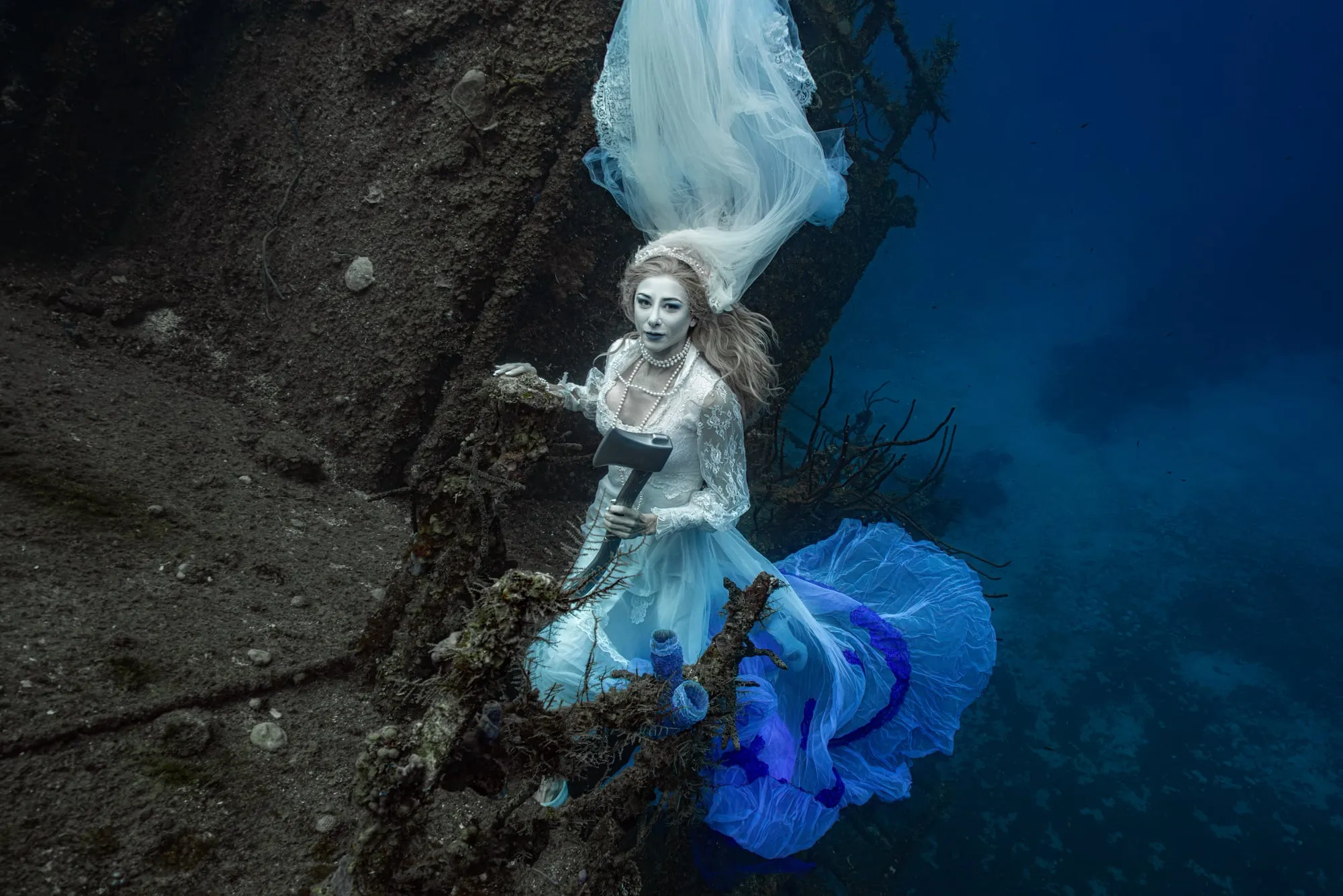 A bride in a white lace gown with a long veil floating above her stands against a dark wreck, a blue and white train drifting beside her.