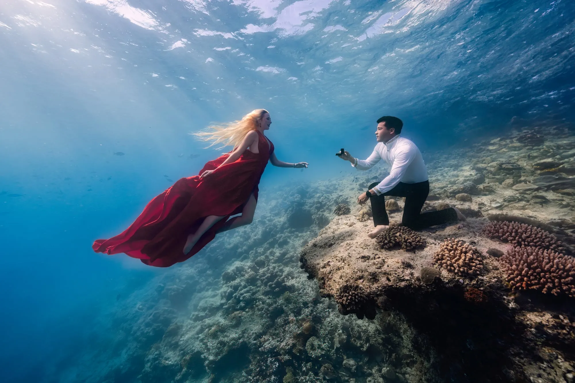 A woman in a flowing red gown floats toward a man kneeling on a coral head and holding out a ring box, sunbeams fanning through the water around them.