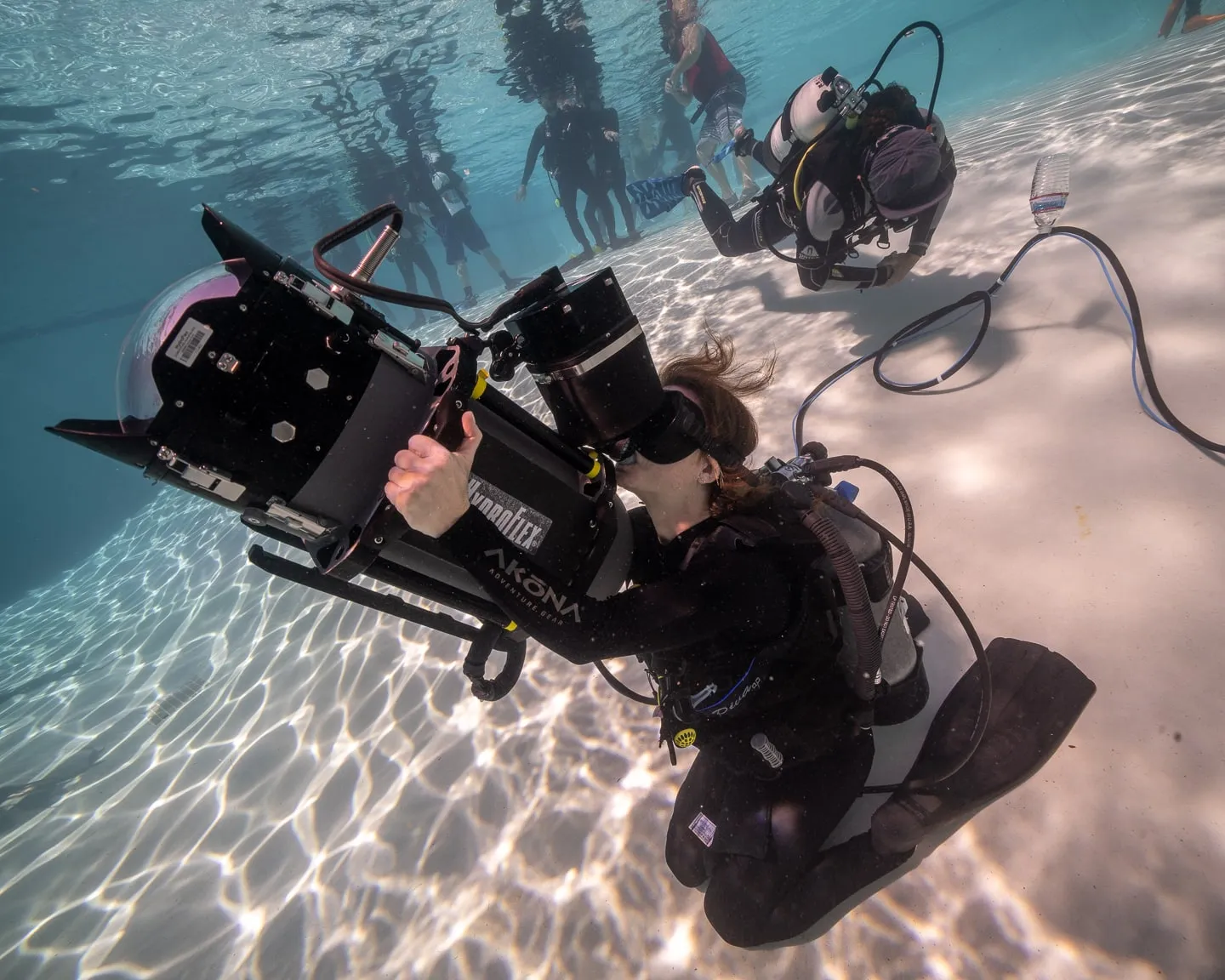 A camera operator in scuba gear on the pool floor steadies a large housed cinema camera, safety divers behind him.