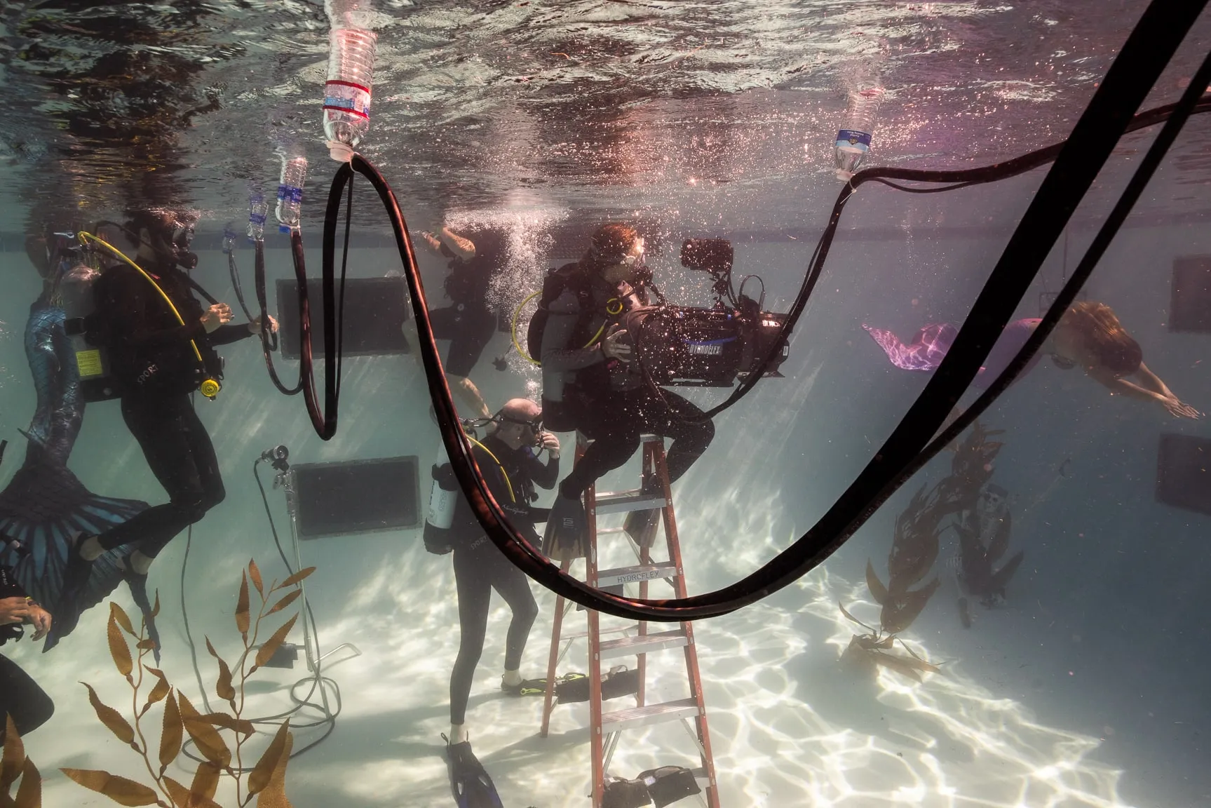 Safety divers in tanks and masks stand around an operator working on a ladder set in a pool, a hose looping overhead.