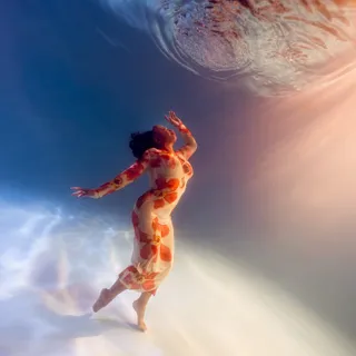 A woman in a red and orange floral dress rises on her toes toward the surface, one arm lifted, sunlight and rays of light streaming across a pale pool floor.