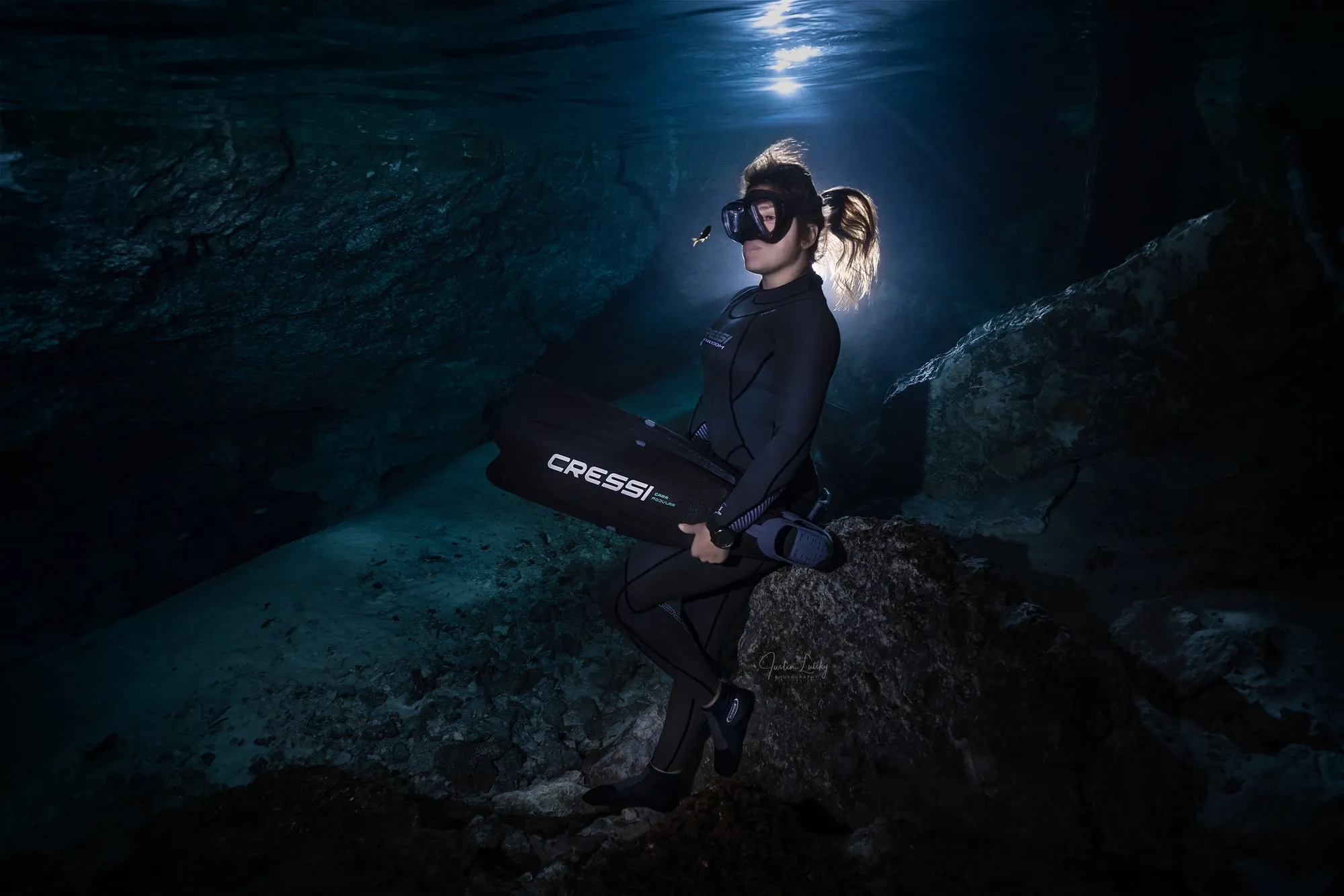 A freediver in a black wetsuit sits on a cave ledge with her fins in her lap, a bright light shining above her in dark water.