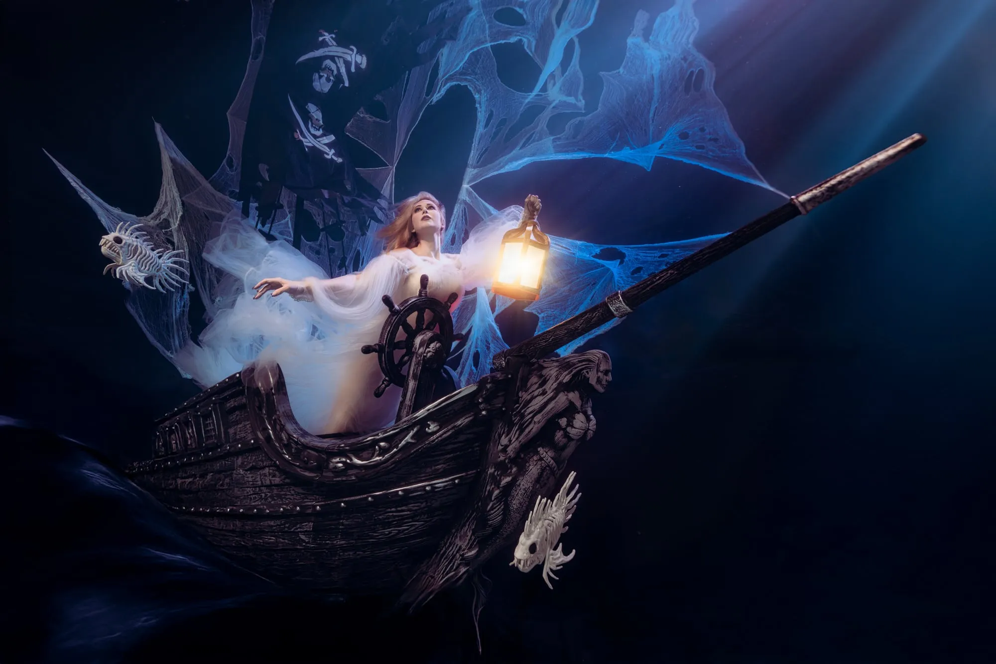 A woman in white tulle lies back on the prow of a black ghost ship, one hand raised toward a glowing lantern, fish skeletons and blue cobwebs drifting around her.