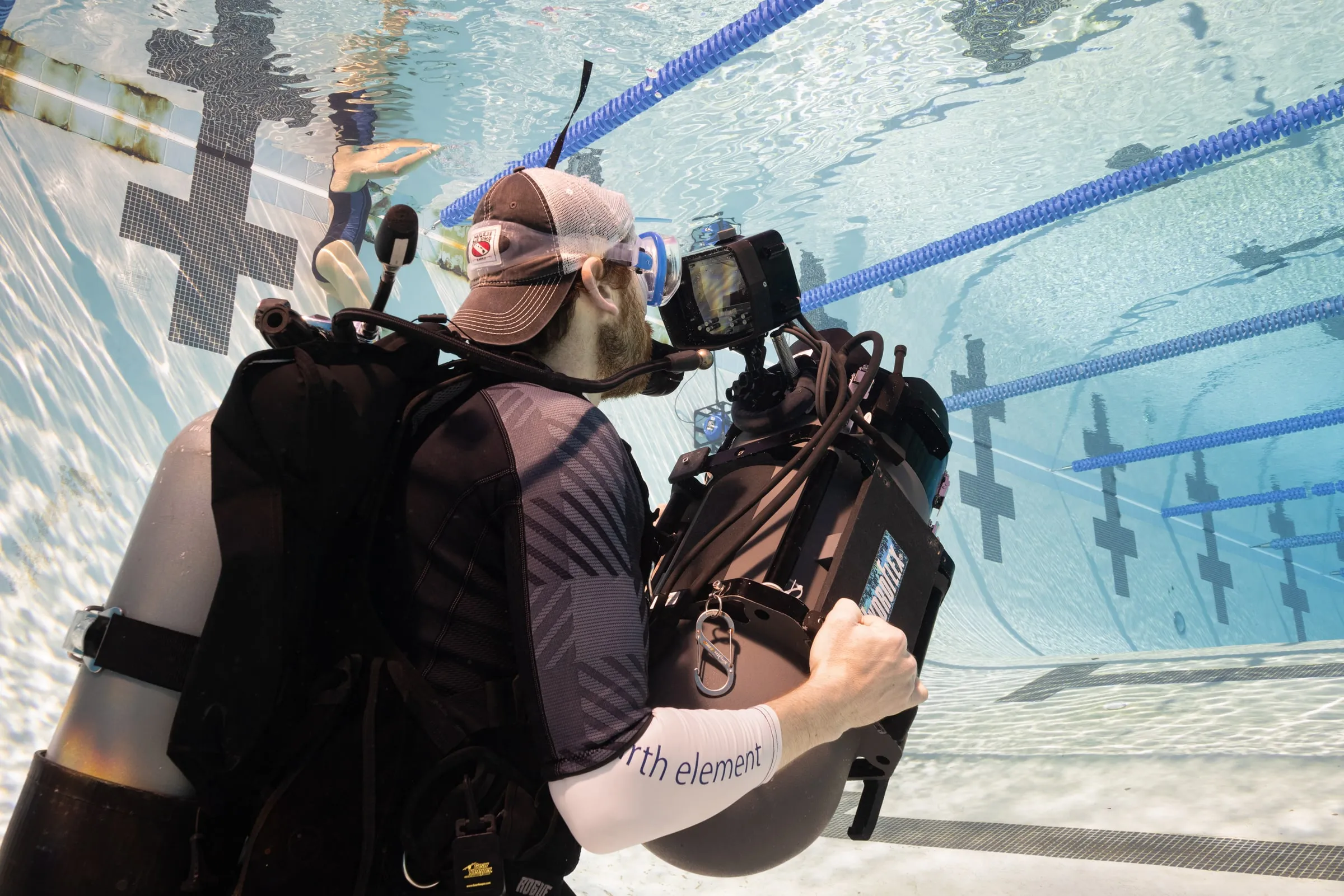 A camera operator in scuba gear holds a cinema camera up to his face in a lap pool, lane lines on the floor behind him.
