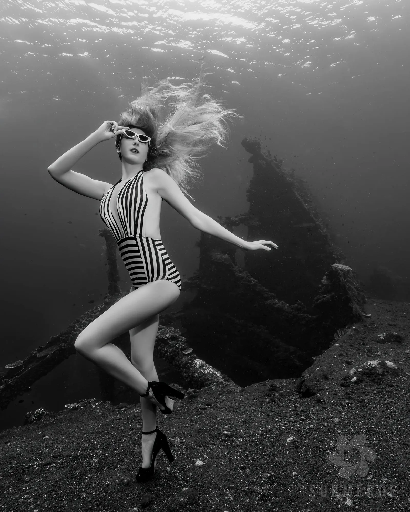 In black and white, a woman in a striped swimsuit, sunglasses and high heels poses with her hair floating up, standing on dark sand beside a wreck.