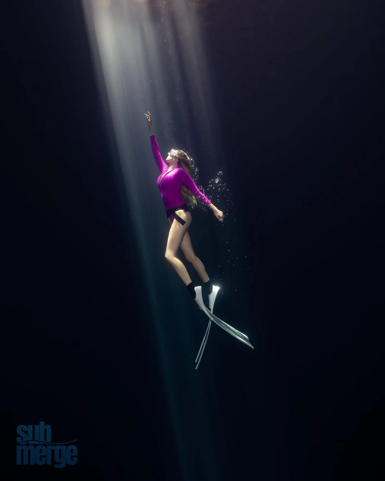 A freediver in a magenta top reaches up as she rises through a shaft of light in black water, long fins trailing below her.