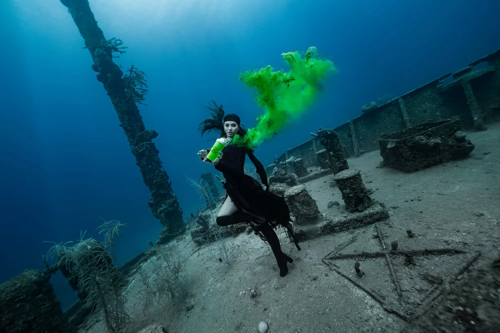A woman in black with a trailing plume of green smoke poses on the deck of a sunken wreck, a mast rising behind her.