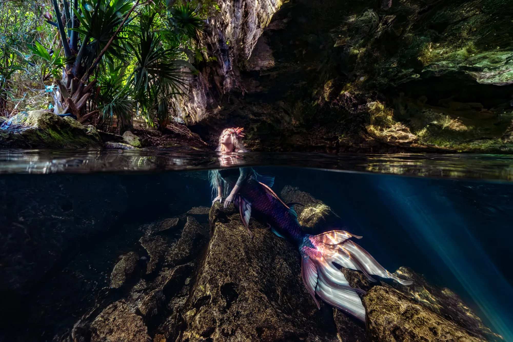 A mermaid with a purple and pearl tail rests on a rock in a dark cenote, her head just above the surface, jungle palms overhead.