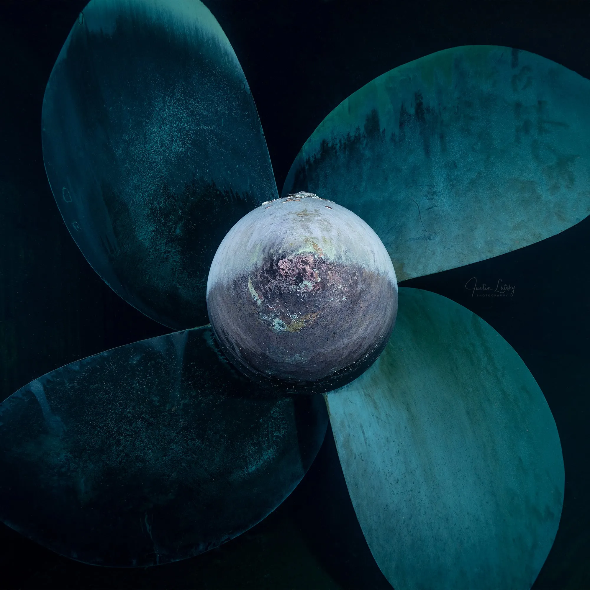 The bronze propeller of a ship seen head-on from below, its hub and four blades lit against dark water.